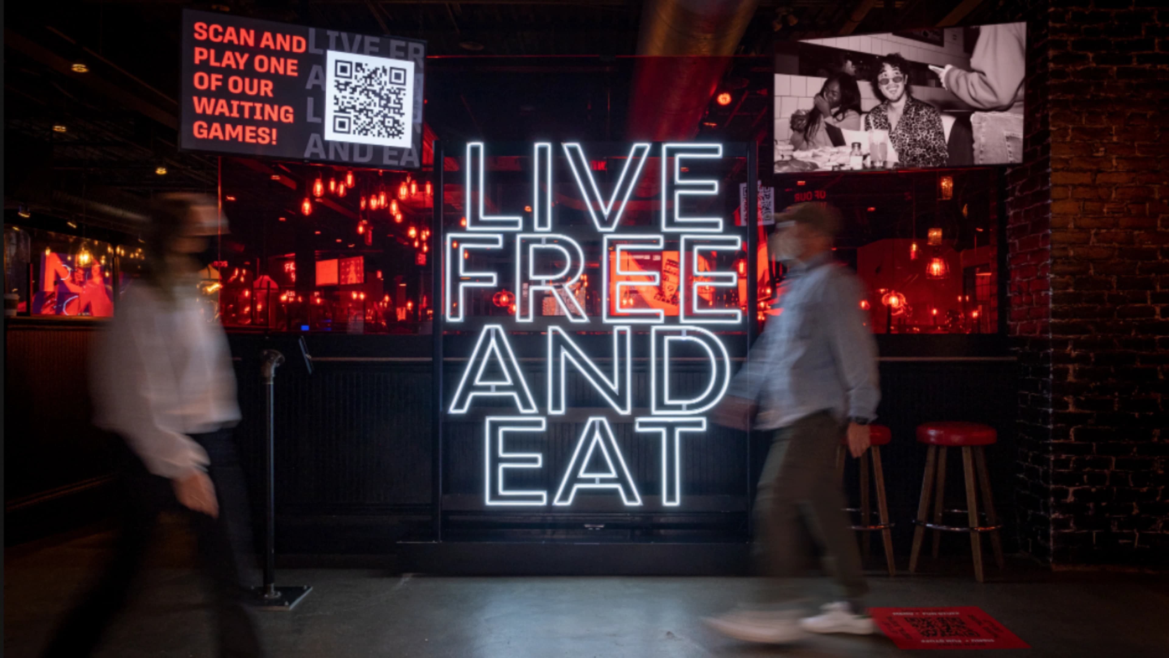 An in-restaurant 'Live Free and Eat' neon sign flanked by a QR-code waiting-game prompt and a photo wall, with guests walking past